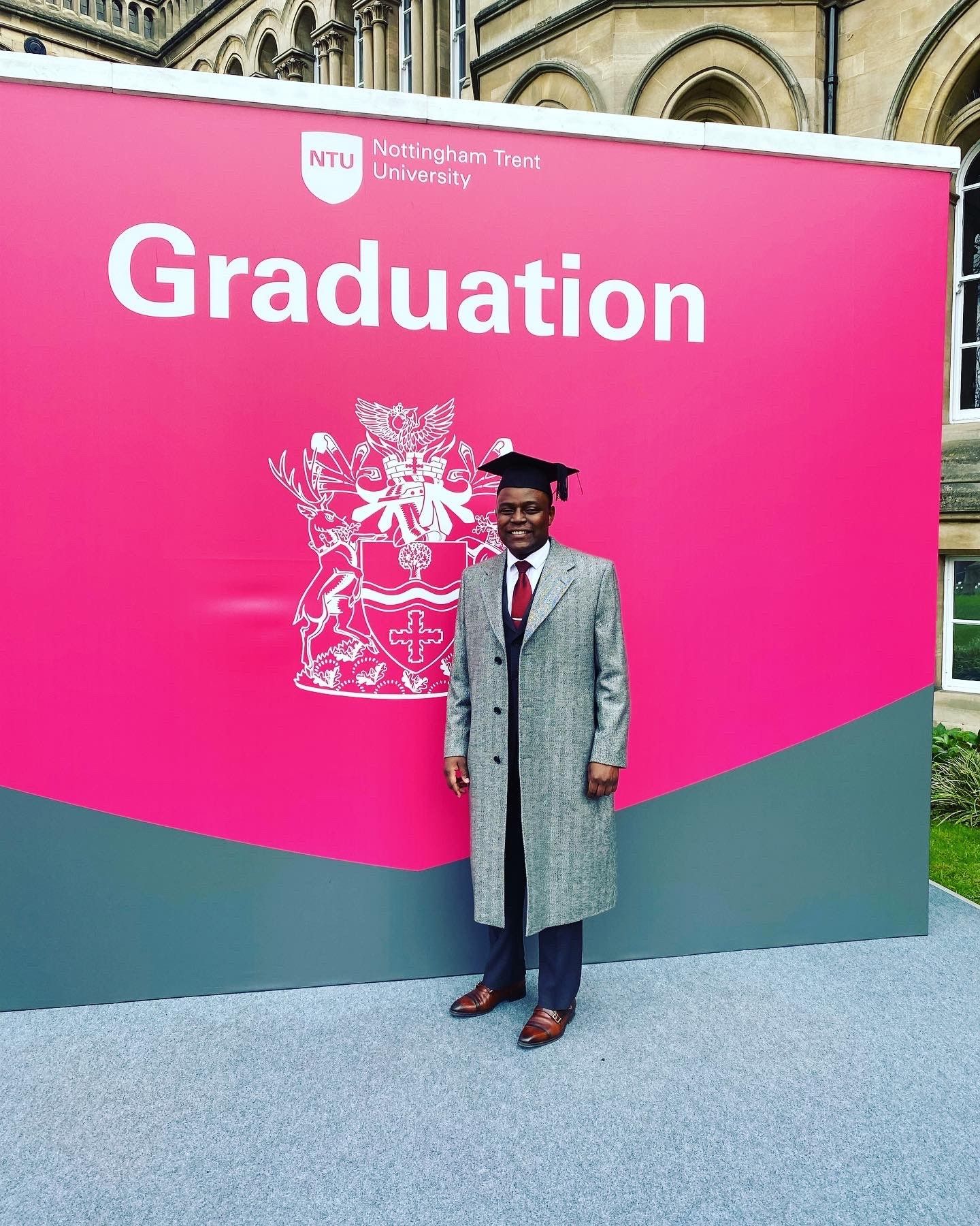 Rodney - who the article is about - stands in front of a big, bright pink banner which says 'Nottingham Trent University Graduation'. He is wearing a graduation mortar-board cap and a long grey jacket and smart shoes. He is smiling.