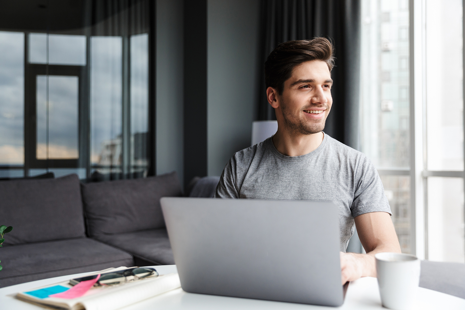 Image of attractive young white man looking out the window with a smile on his face, with a laptop in front of him.