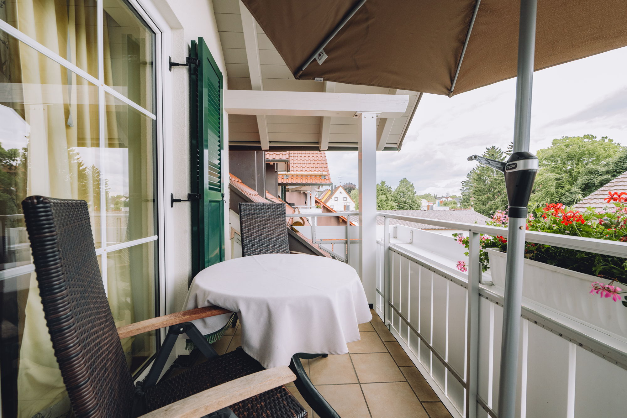 Ein Balkon mit Sonnenschirm, weißem Tisch, verstellbaren Stühlen und Blumenkasten.