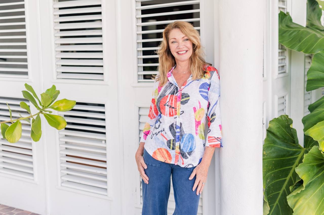 A woman wears a colorful patterned top with blue hemmed pants and is leaning on a beach porch.