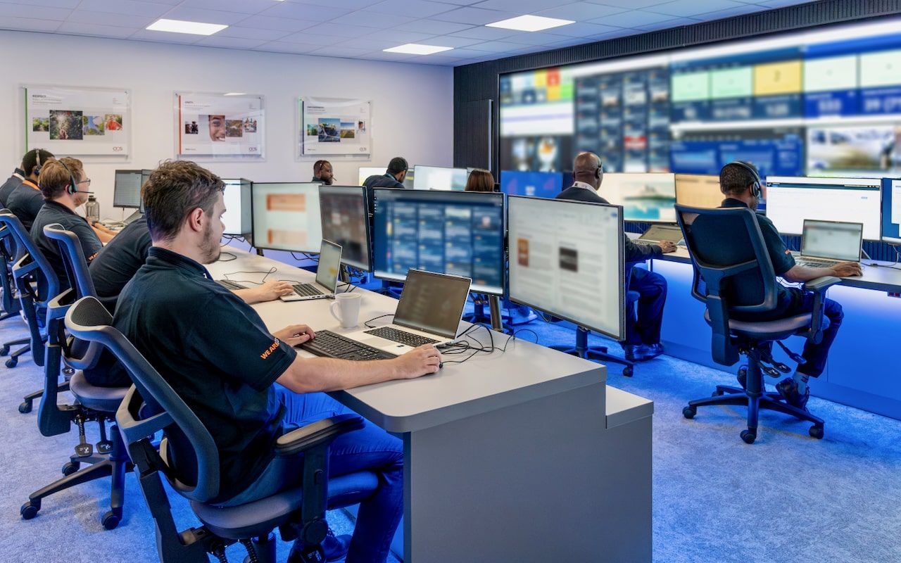 Several people sit at desks with multiple monitors in a control room, working and monitoring digital displays. Large screens cover the front wall, showing various data and information. The room is brightly lit and organized.