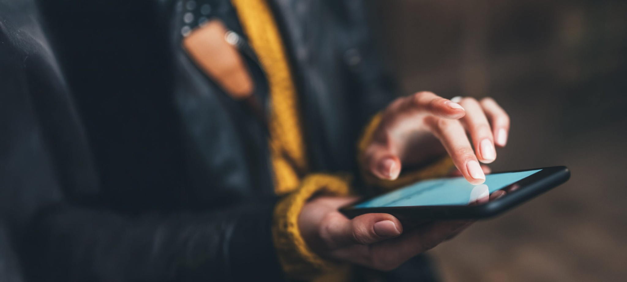 Close-up of a person wearing a dark jacket and yellow scarf using a smartphone, with one hand holding the device and the other hand touching the screen. The background is blurred.