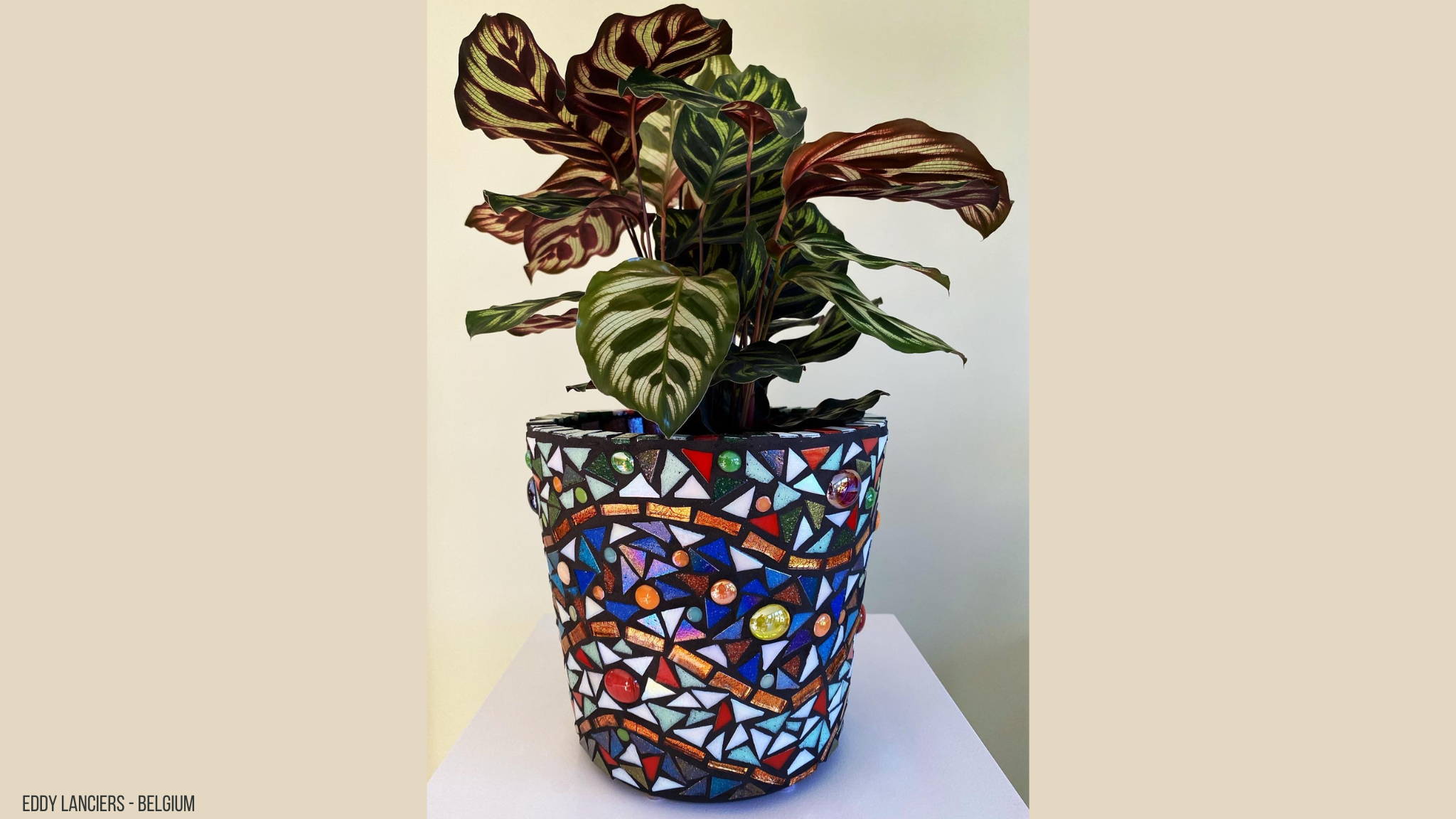 Een plant in een bloempot die bezet is met glasmozaiek en glasdruppels. Er zitten blauw, wit en rode tinten in.