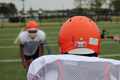Football players in uniform with orange helmets and white jerseys. The player in the foreground is turned to face away from the camera, with a Tozuda Impact Indicator visible on the back of the helmet.