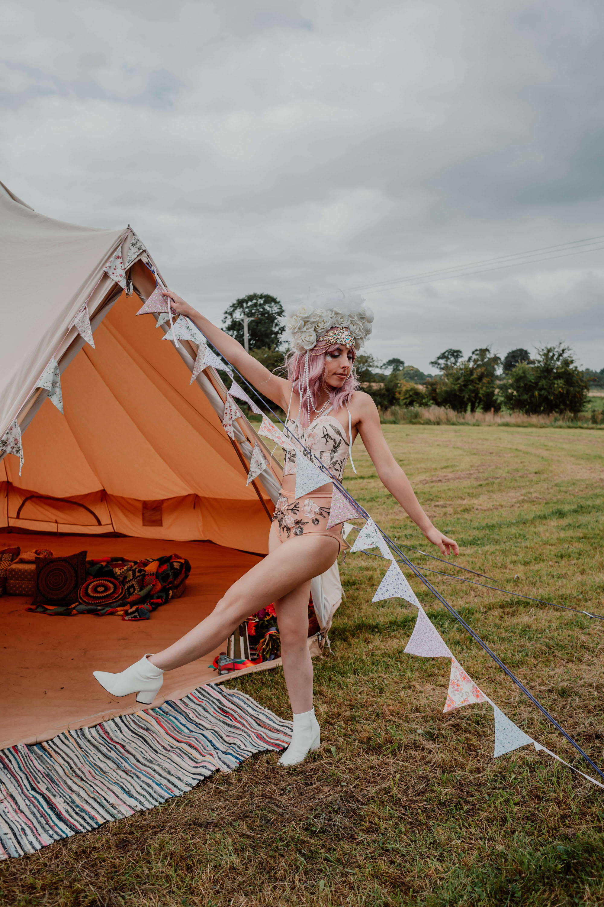 Image of a bride at a festival. She is wearing an embroidered modern bridal outfit, white head piece instead of a veil, and has biodegradable glitter on her face. She's standing in front of a bell tent which looks warm and inviting, and has bunting and colourful decor.