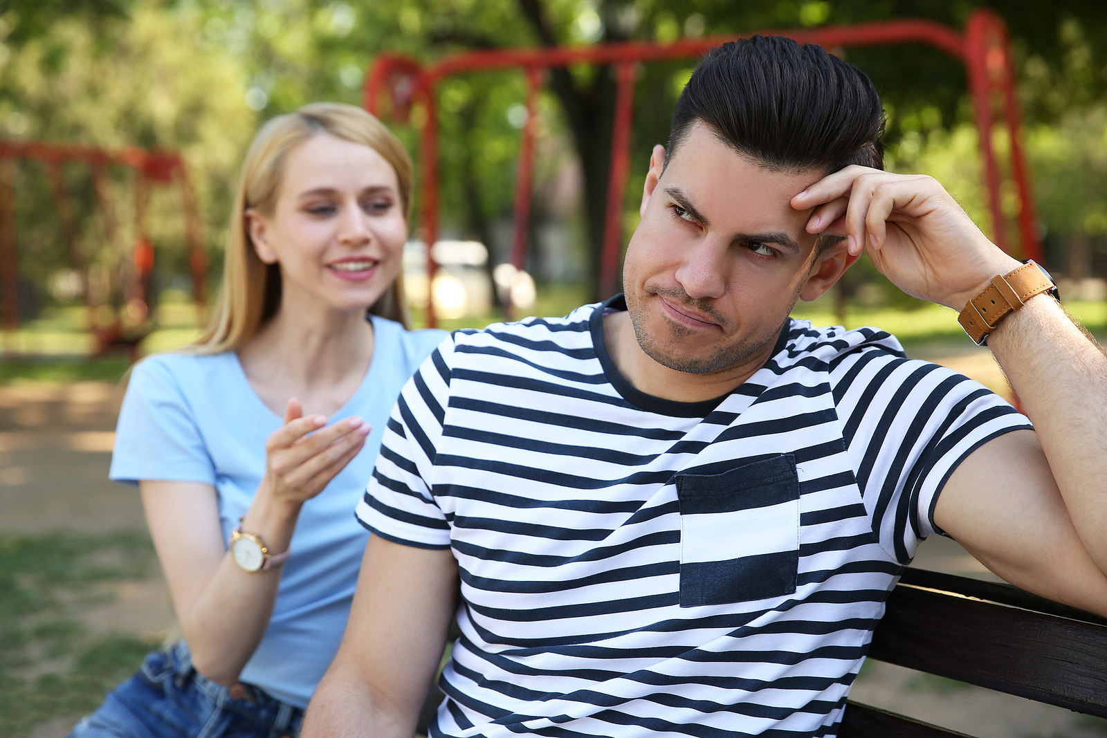 Image of a young man with a striped shirt, looking away with an annoyed look while his date talks to him while they sit on a bench.