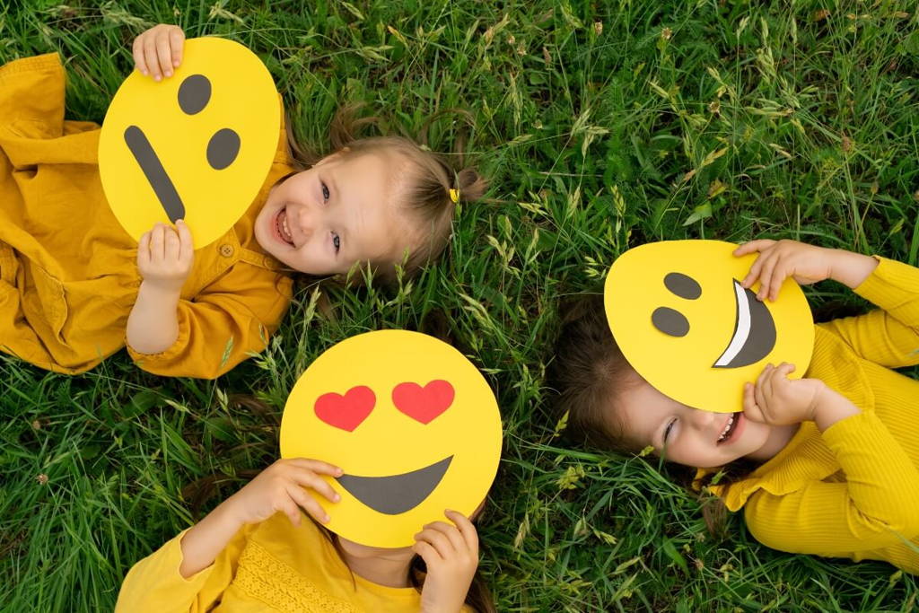 Three young children lying on grass, each holding a large yellow emoji face cutout showing different emotions—neutral, heart eyes, and smiling—while wearing bright yellow clothing and laughing together.