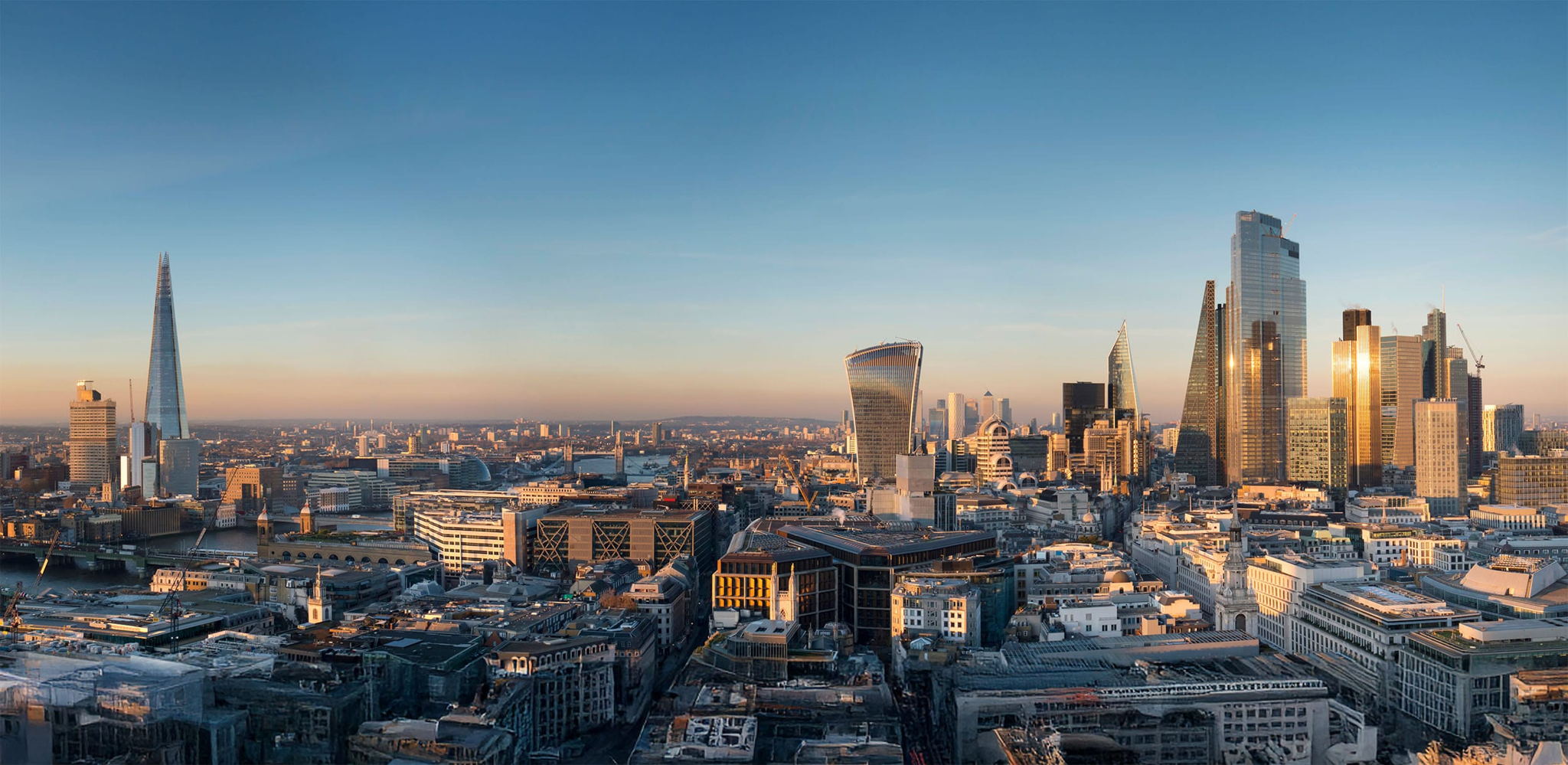 A panoramic view of London’s skyline at sunset, featuring modern skyscrapers like The Shard and the Walkie Talkie building, with the River Thames and historic city buildings visible under a clear blue sky.