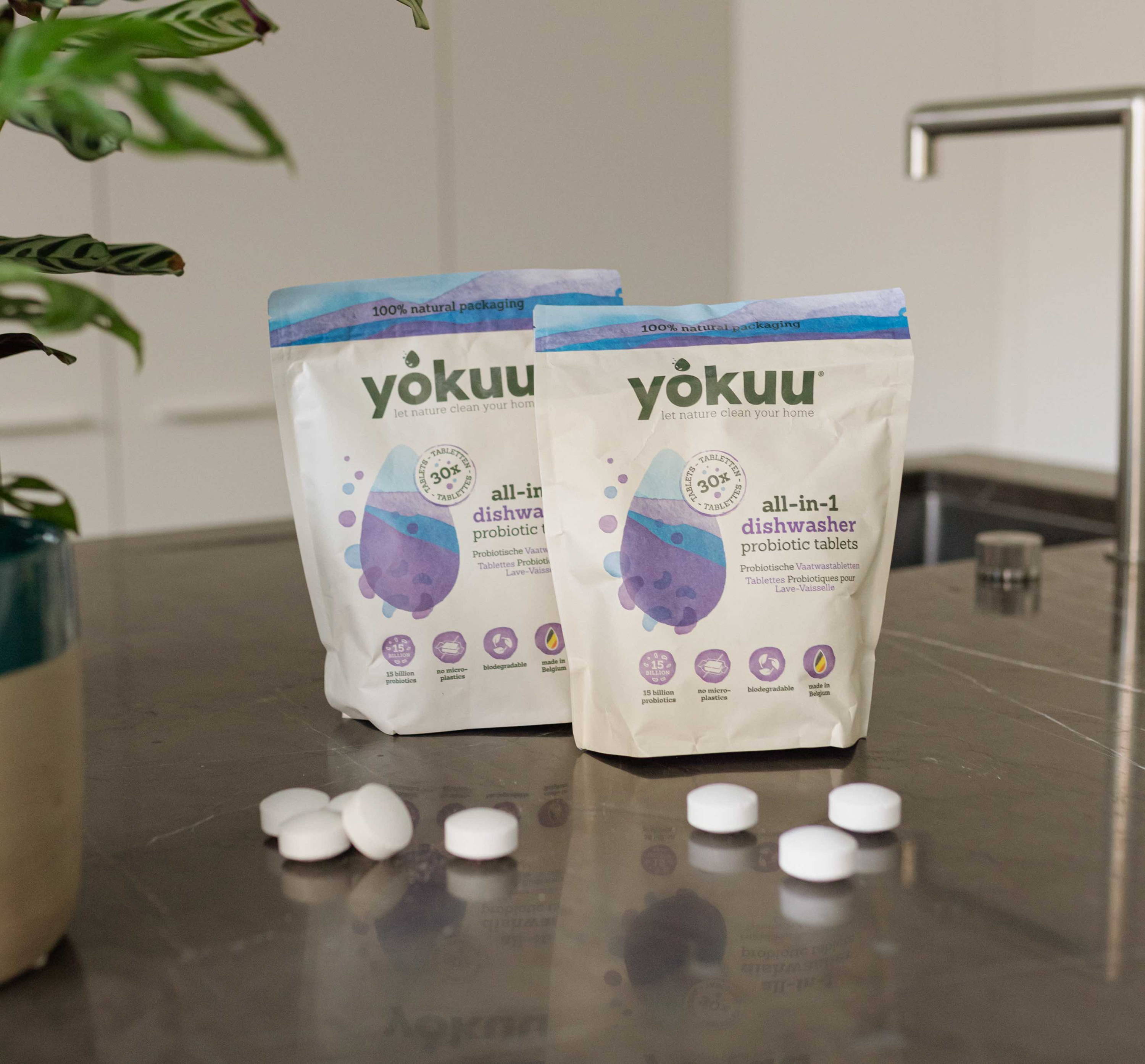 Two Yokuu dishwasher tablets packages are placed alongside plastic-free tablets on a marble surface with a plant in the foreground..