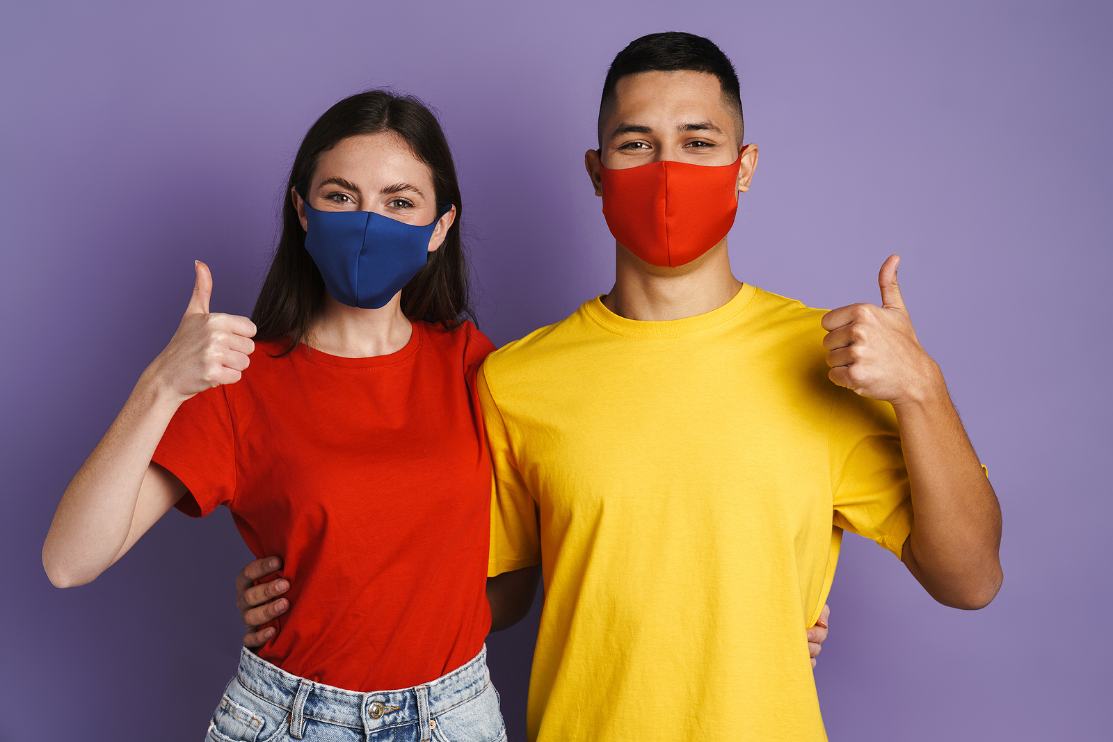 A man and woman smile with face masks on while giving a thumbs up.