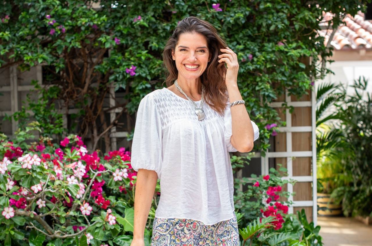 A woman in a white sleeve blouse and patterned pants stands in front of a flowering garden.