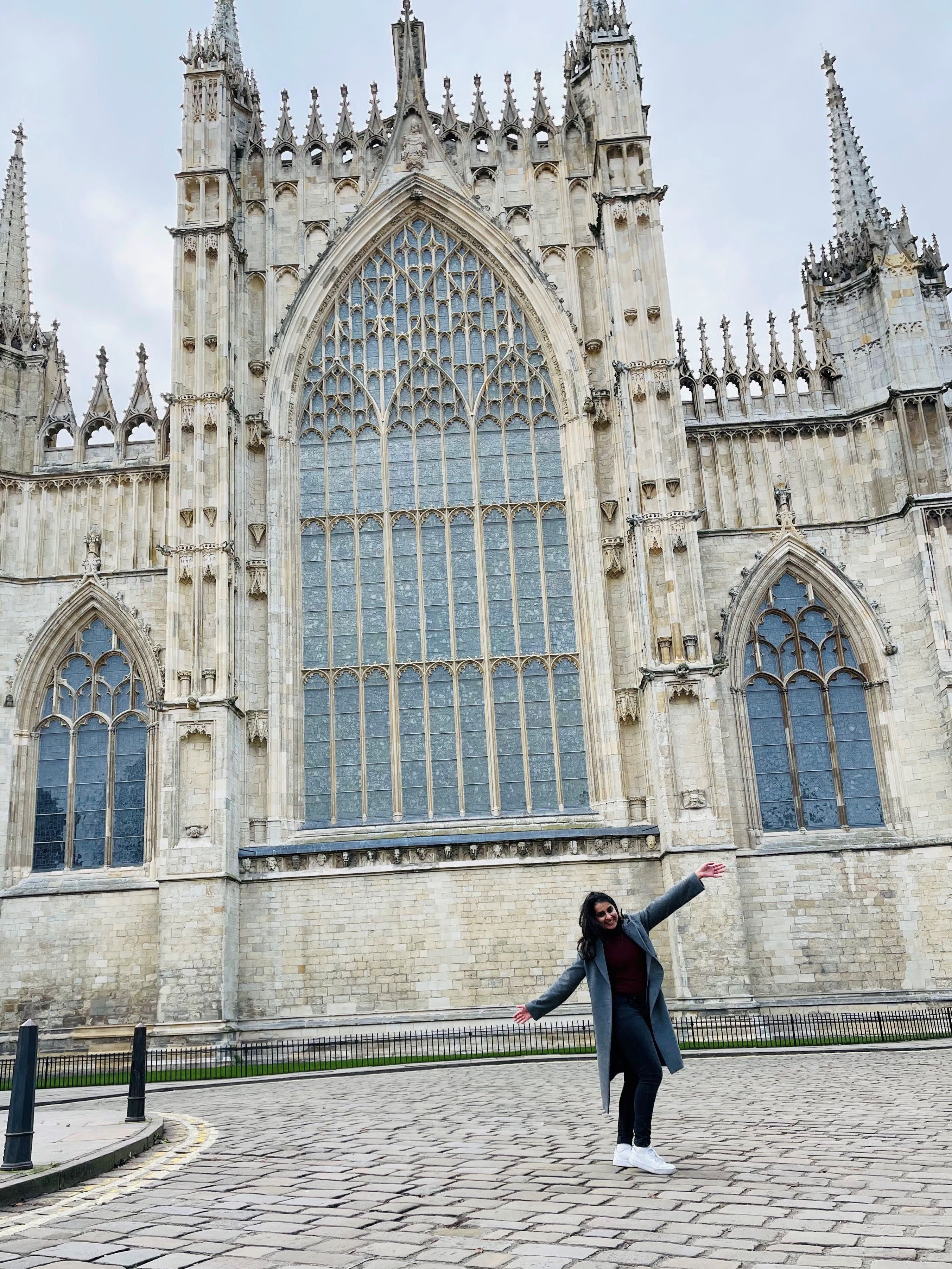 International student in front of York Minster on a grey day. She is posing with her arms wide, in a diagonal fashion, and is smiling.