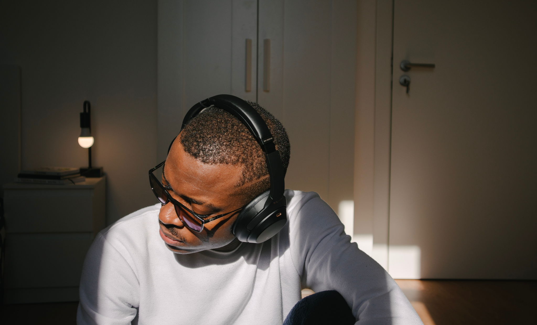 A black man wearing headphones and listening to music in his bedroom looks down and to the side. He is sitting by the window.
