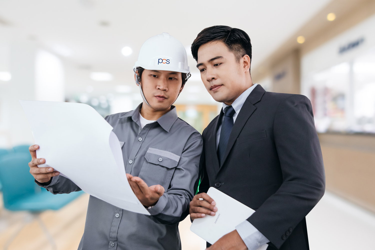 Two men standing indoors, one wearing a white construction helmet and gray shirt holding blueprints, the other in a dark suit holding documents. They are looking at the papers together in a professional setting.