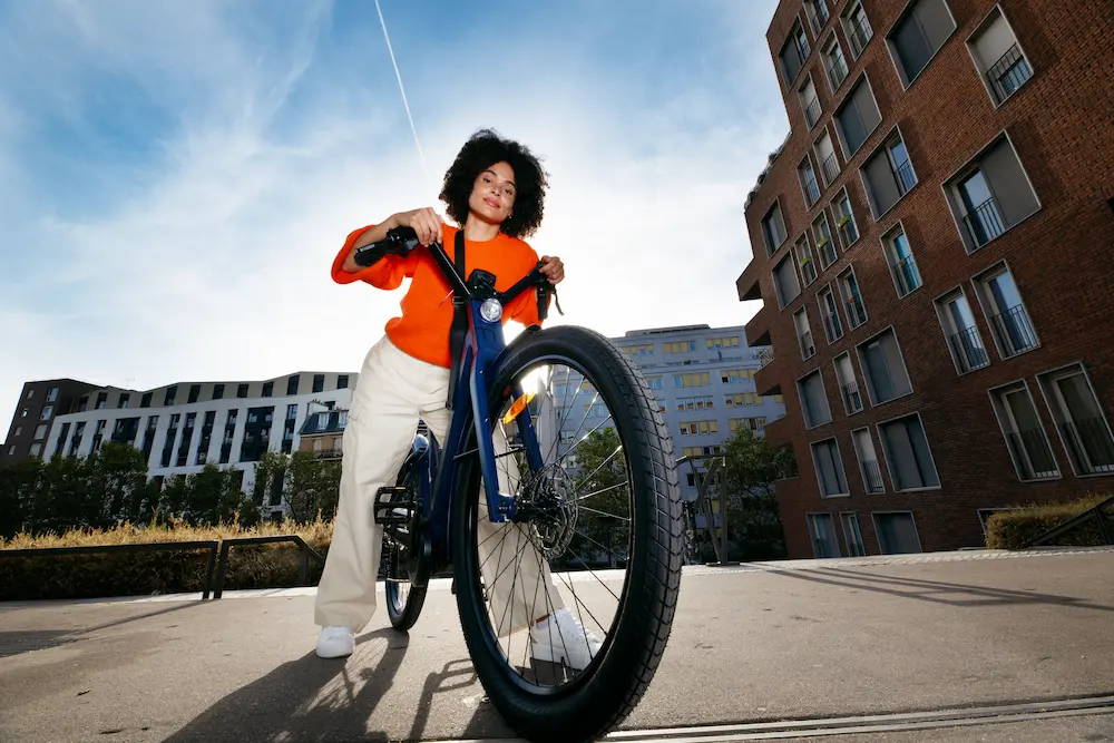 Frau in orangefarbenem Oberteil und heller Hose lehnt sich auf den Lenker ihres Fahrrads. Sie steht auf Asphaltboden. Im Hintergrund sind moderne Gebäude. 