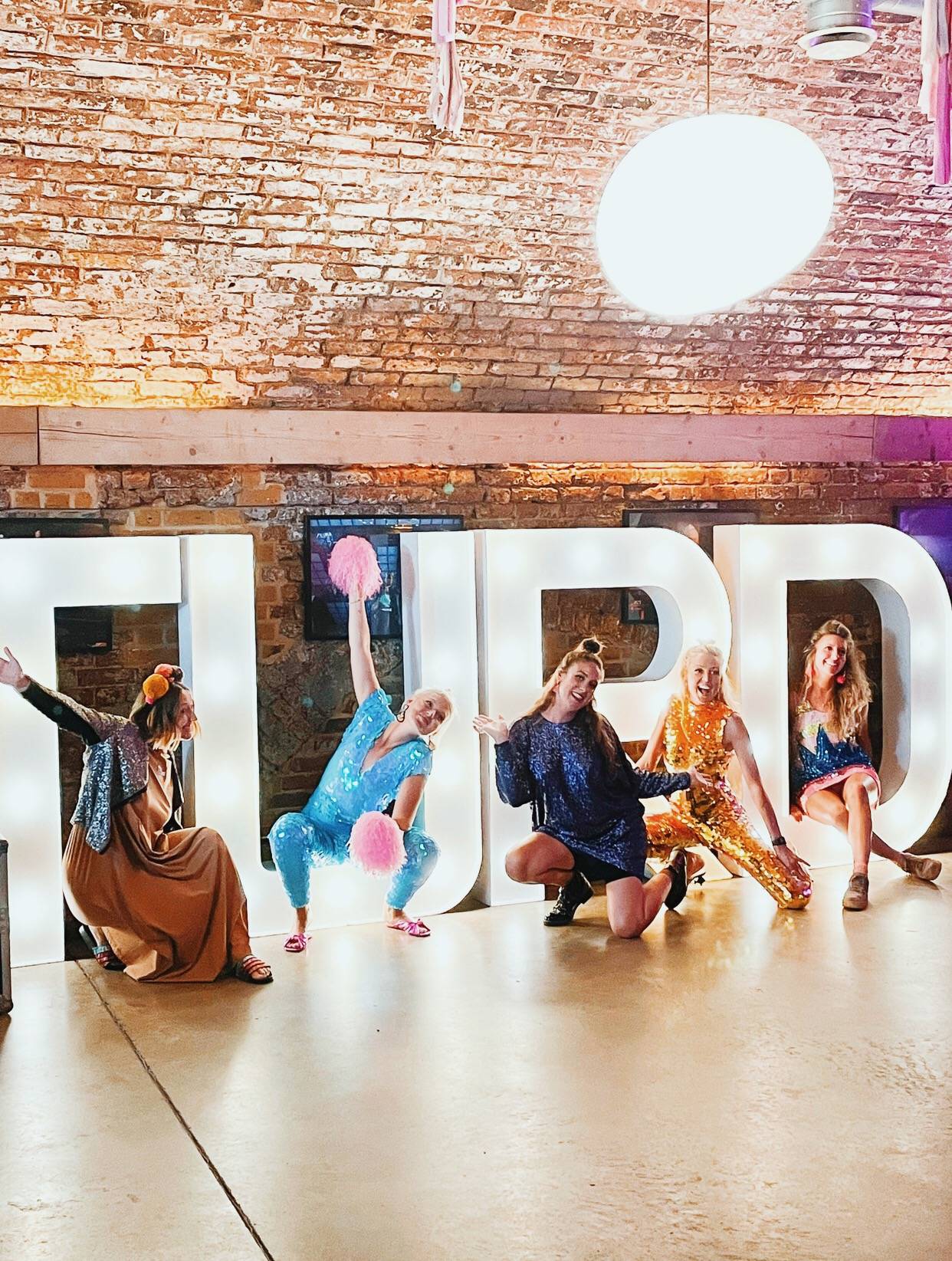 Image shows women in sparkly outfits with biodegradable glitter on their faces posing in front of giant white letters that spell "turd".