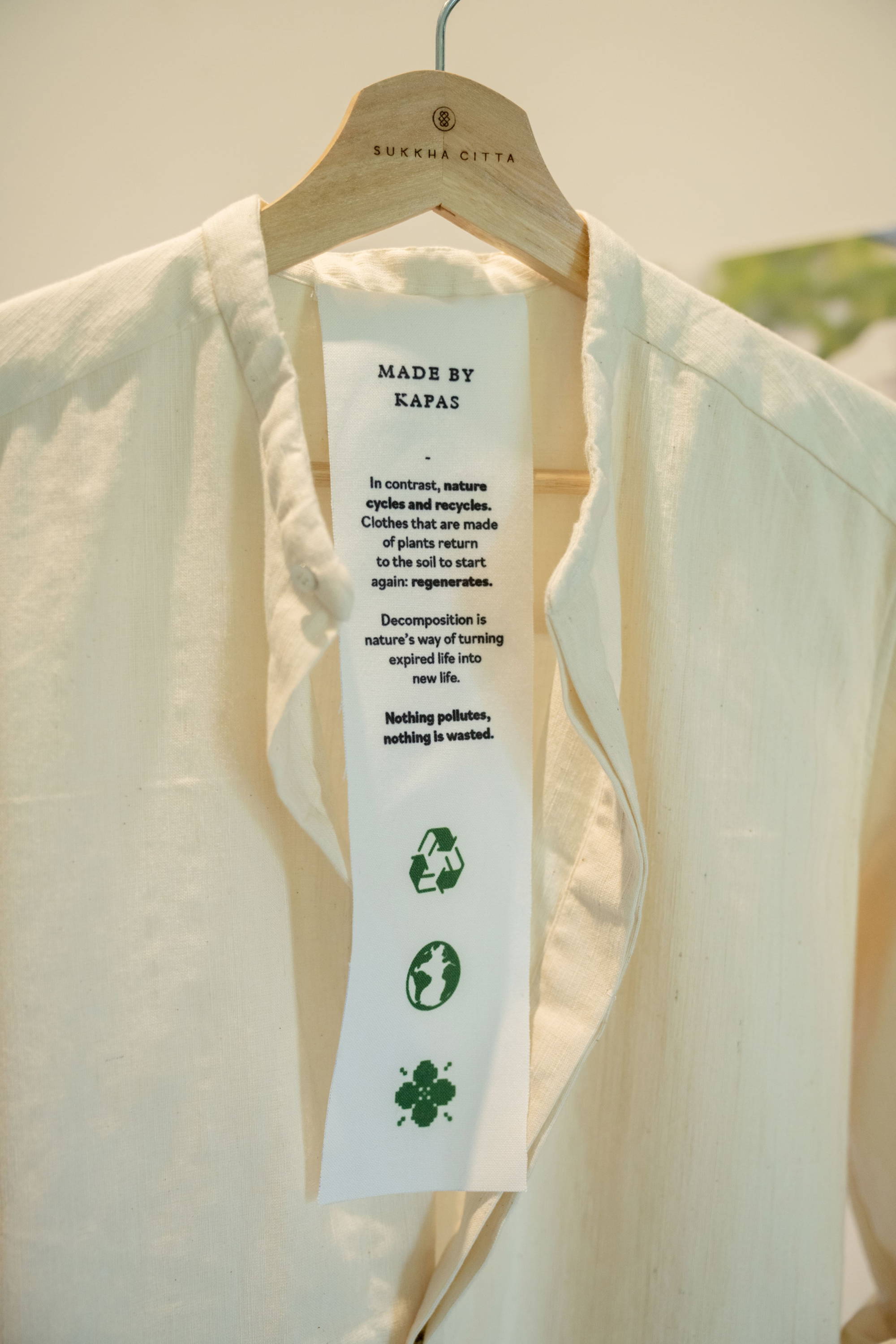 KAPAS Installation by SukkhaCitta in John Hardy Boutique & Gallery at Seminyak. Hangtag from a white dress shirt made of regenerative cotton, addressing its environmental impact. Text reads: In contrast, nature cycles and recycles. Clothes that are made of plants return to the soil to start again: regenerates. Decomposition is nature's way of turning expired life into new life. Nothing pollutes, nothing is wasted.