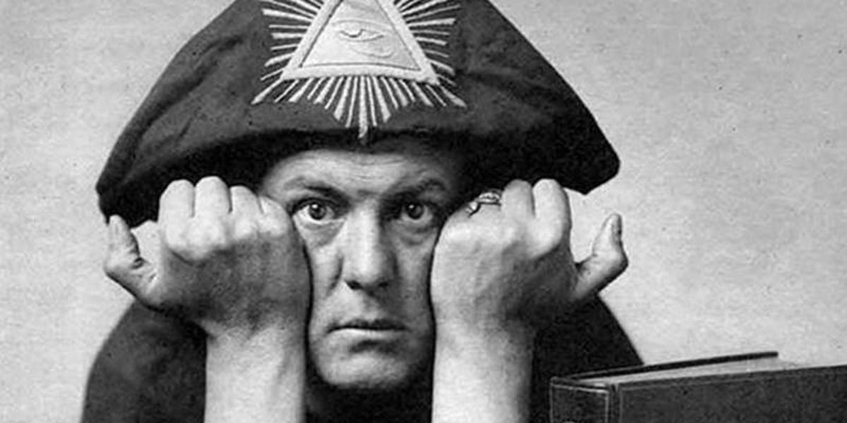 Black and white image of Aleister Crowley wearing his ceremonial garb. He has his hands in fists against his cheeks.