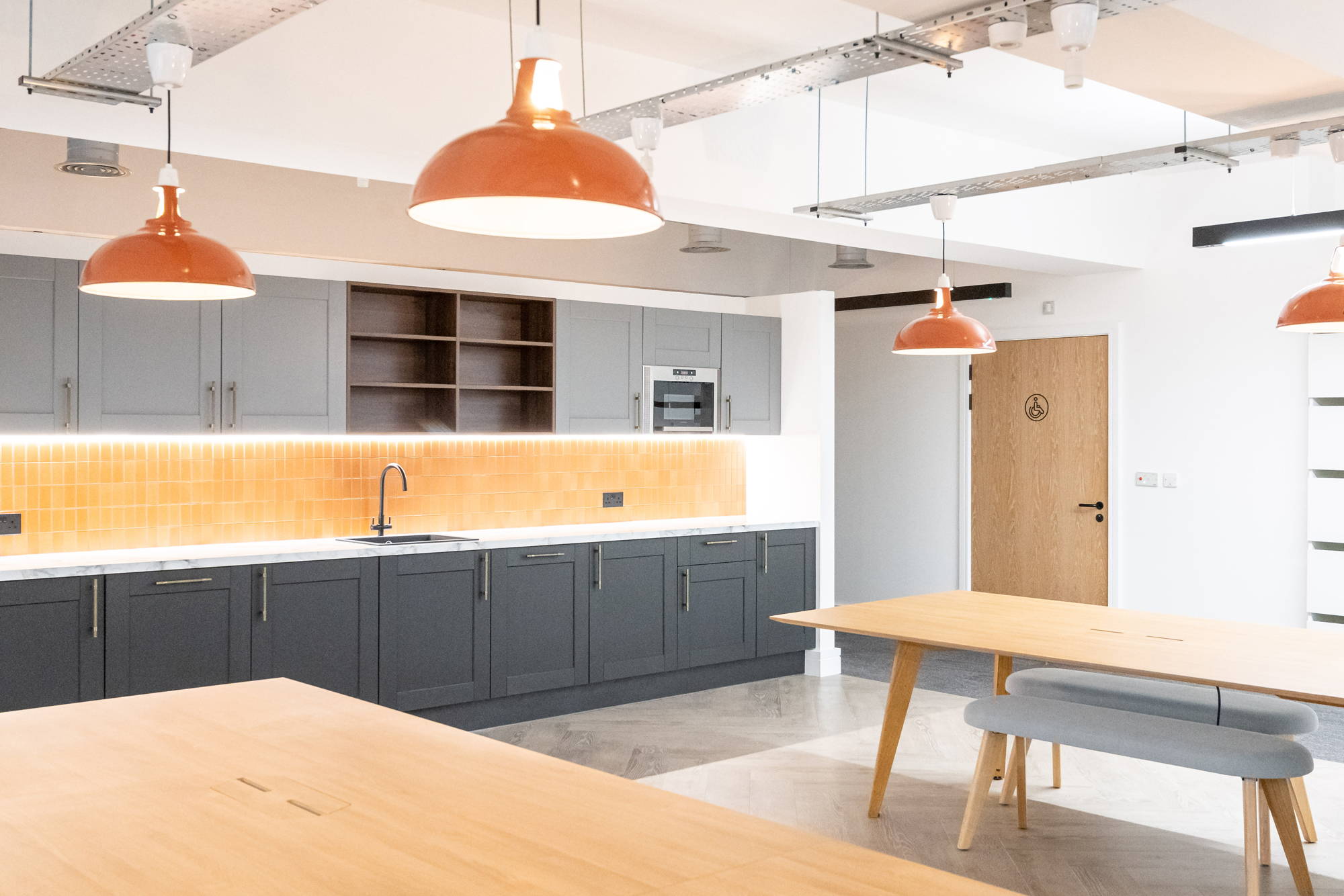 4 Large Terracotta Lampshades hang above 2 large wooden tables in a shared commercial workspace. Next to the tables is a kitchenette with black and grey cupboard doors.