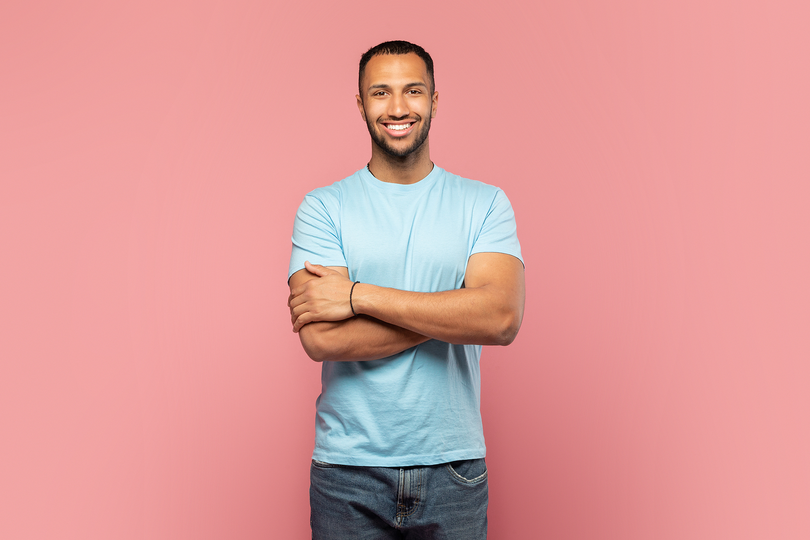 Portrait of happy african american guy standing with folded arms and smiling, posing over pink background.