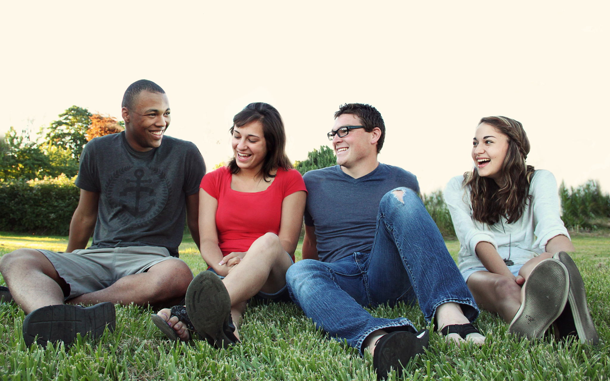 An group of 4 friends of different ethnicities sit together in the grass laughing together.