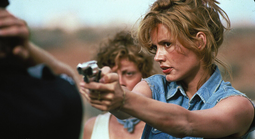 Louise stands behind Thelma as she points her gun towards an officer with very tense expression.