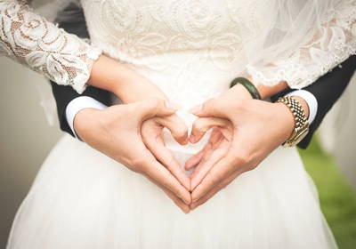 Jeune couple en habits de mariage qui forment un coeur superposé avec leurs quatre mains sur le ventre de la femme.