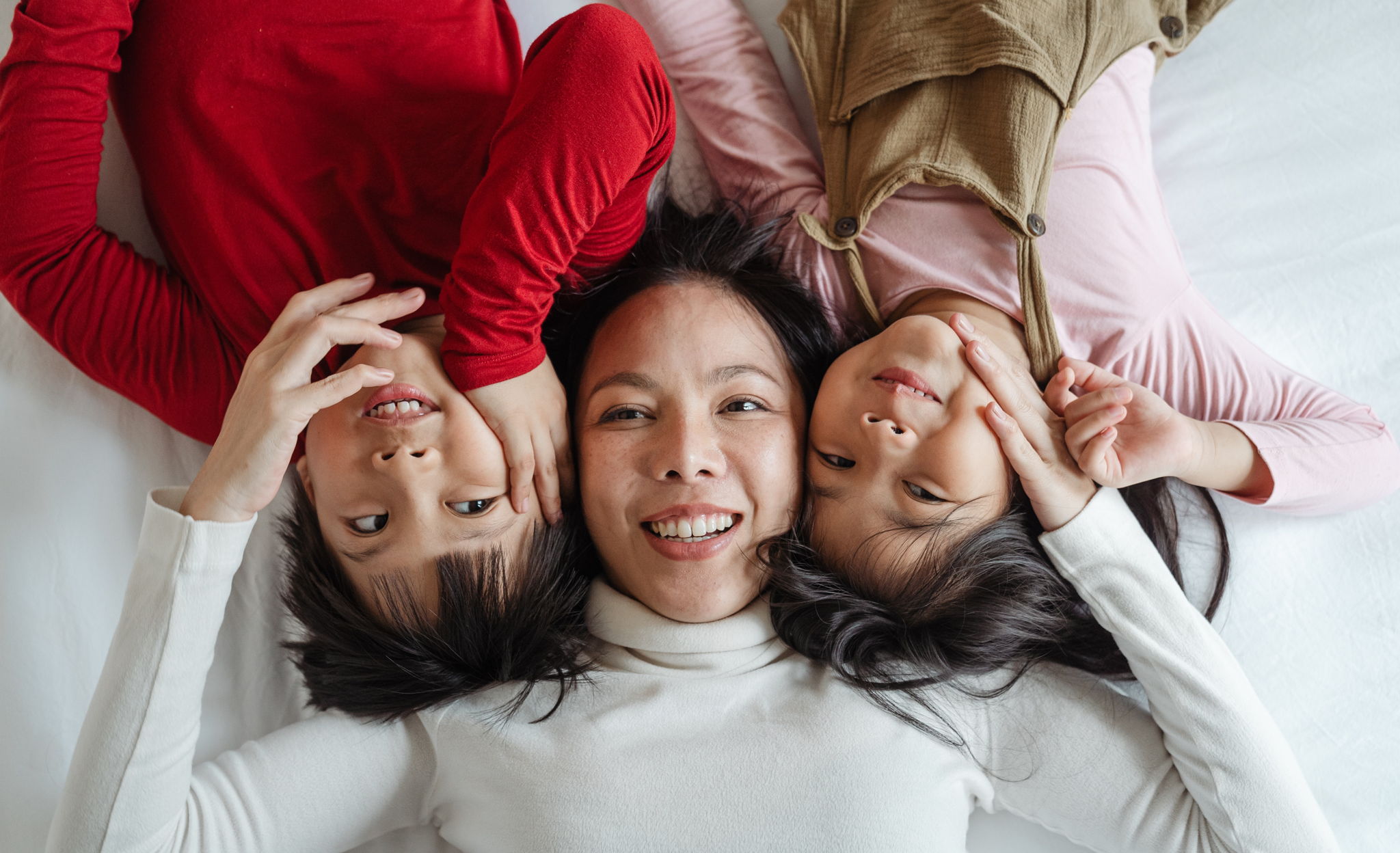 An Asian mom and her children, laying on top of a white bed smiling to eachother.