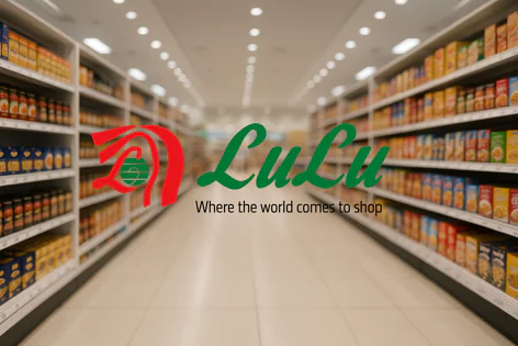Red and green LuLu logo and slogan "Where the world comes to shop" over a blurred supermarket aisle filled with groceries on shelves.