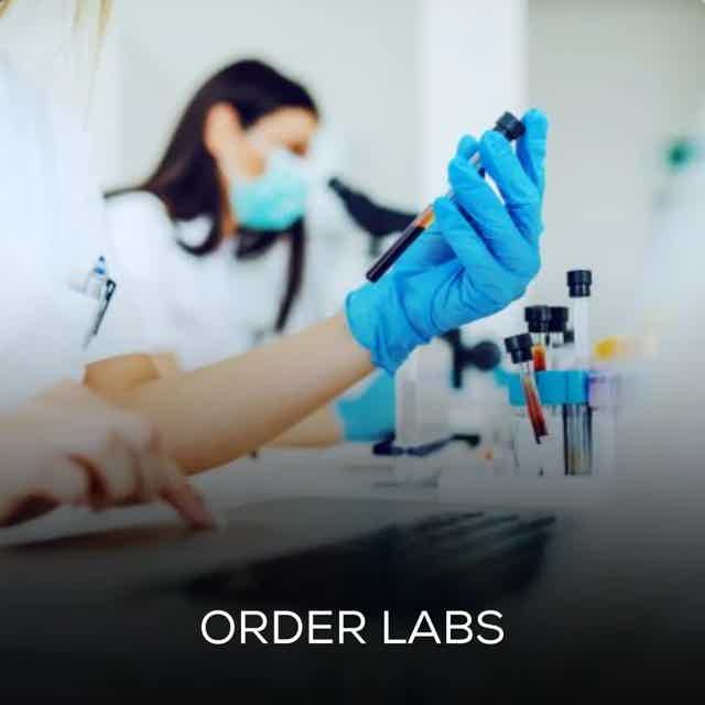 Lab tech wearing blue gloves holds a sample tube in a lab with others and a microscope in a blurred background.