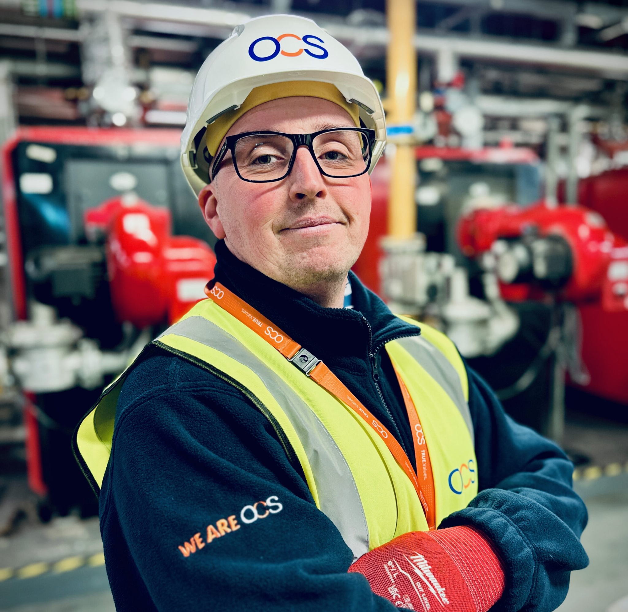 A confident worker in a hard hat, safety glasses, and high-visibility vest stands with arms crossed in an industrial setting. The uniform and helmet display the OCS logo. Pipes and machinery are visible in the background.
