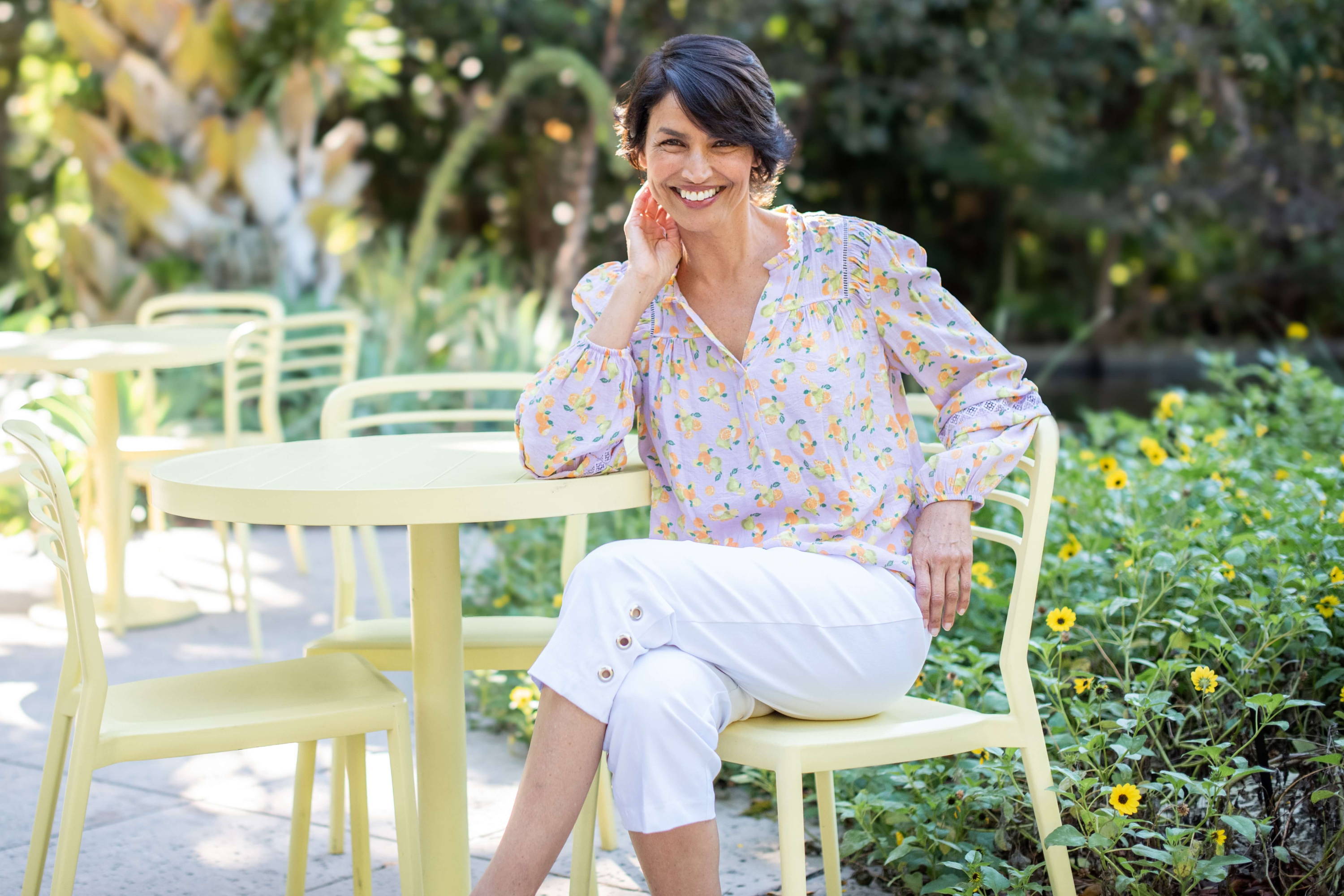 A woman in a floral blouse and white crop pants sits at a bistro table on an outdoor garden patio