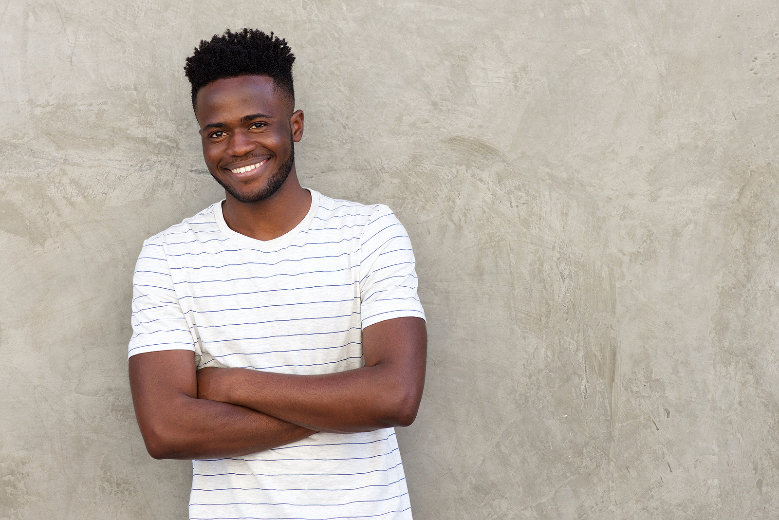 An attractive black guy wearing a striped shirt has his arms crossed with a confident smile, leaning against a wall.