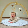 Baby smiling and laying on a soft mat while a wooden arch with mobile plush toys is above her.