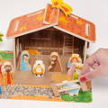 Hand placing a wooden fence piece onto the Montessori Nativity Scene set board, with other figures and the barn visible in the background.
