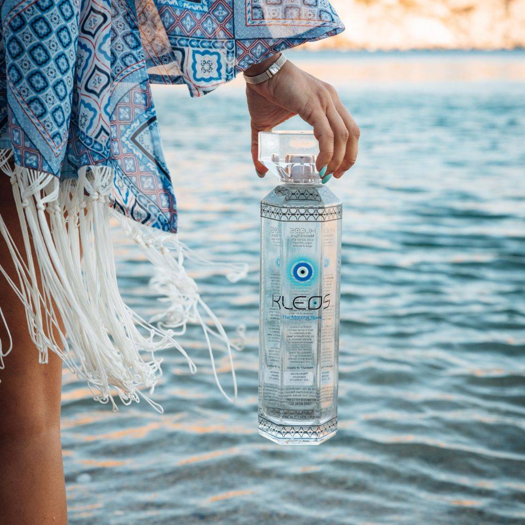 Woman holding bottle of Kleos Mastiha Spirits on a beach - click to shop products