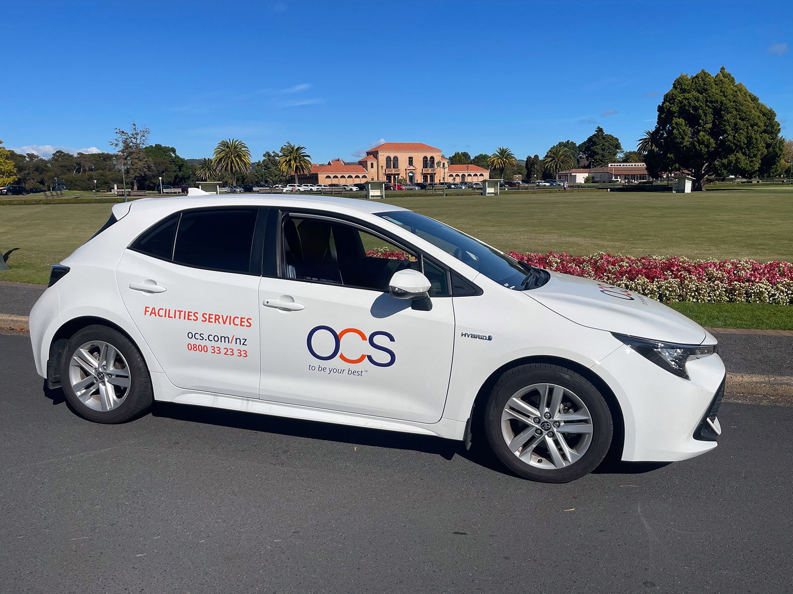 A white OCS Facilities Services hybrid car is parked on a road beside a grassy park with palm trees and a large building in the background. The car displays the OCS logo and contact details on its side.