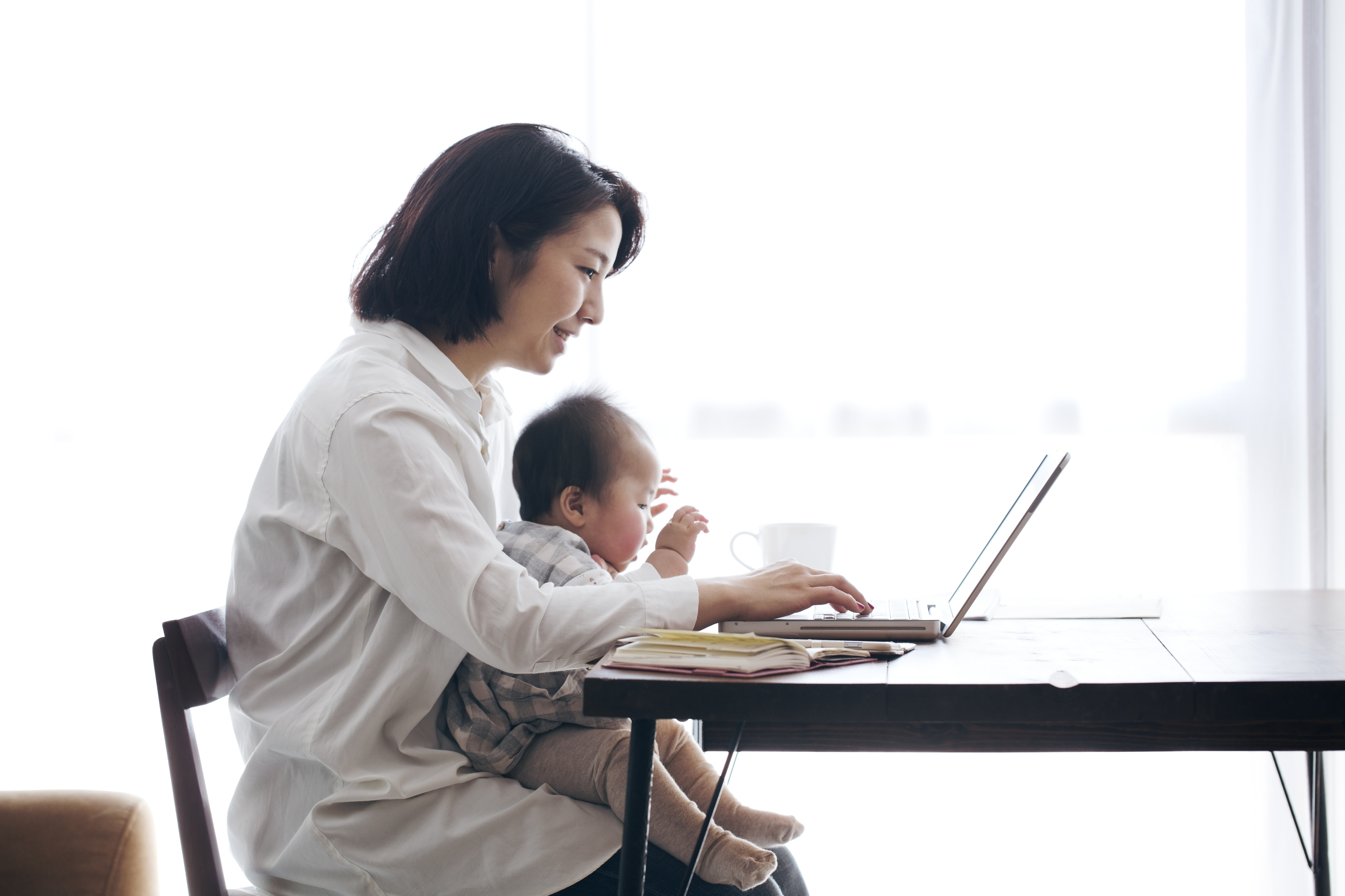Working mother smiles, holding baby in lap and looking at laptop