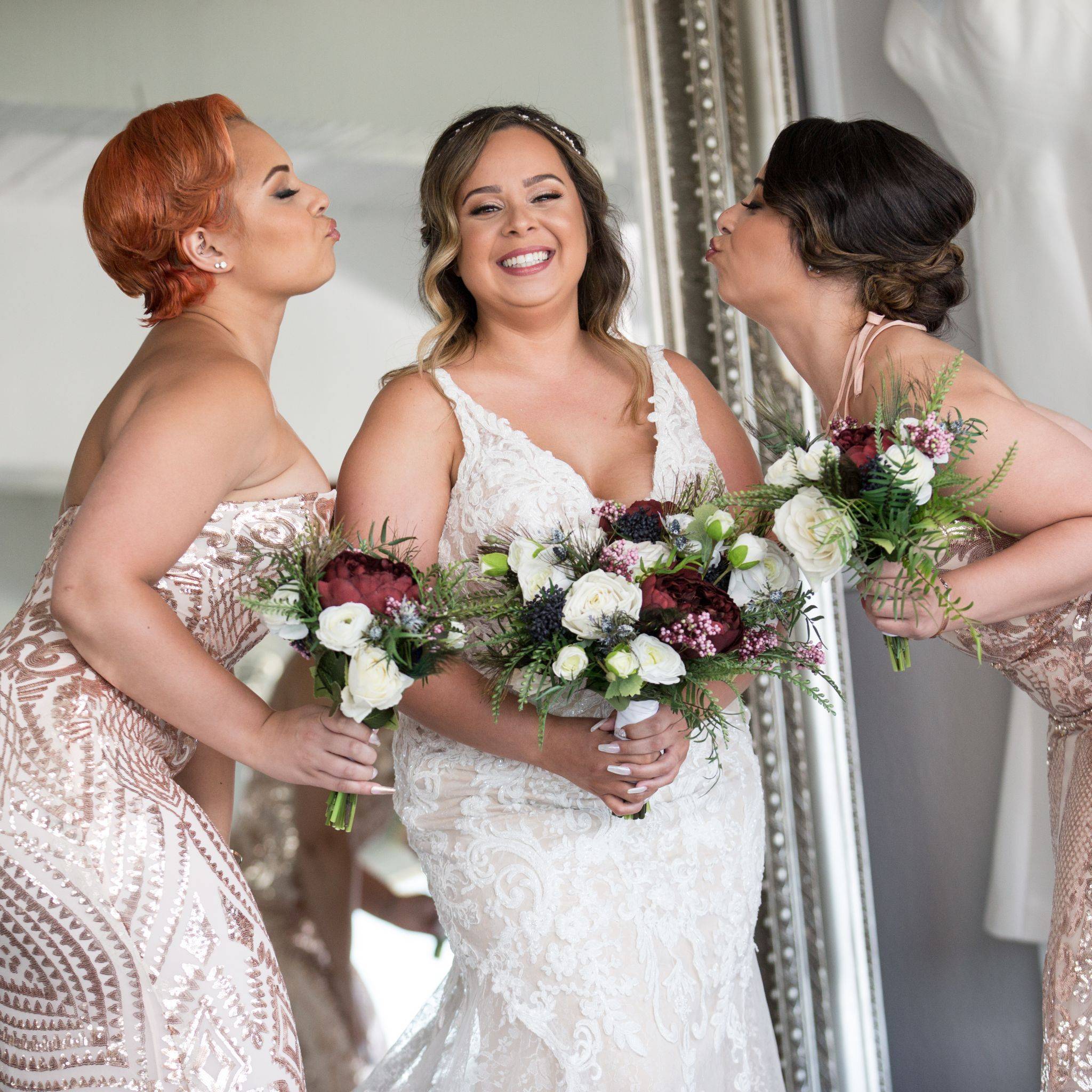A bride holding her bouquet made of maroon, burgundy, blue and white flowers receives air kisses from her bridesmaids holding smaller bouquets made of the same high quality faux flowers