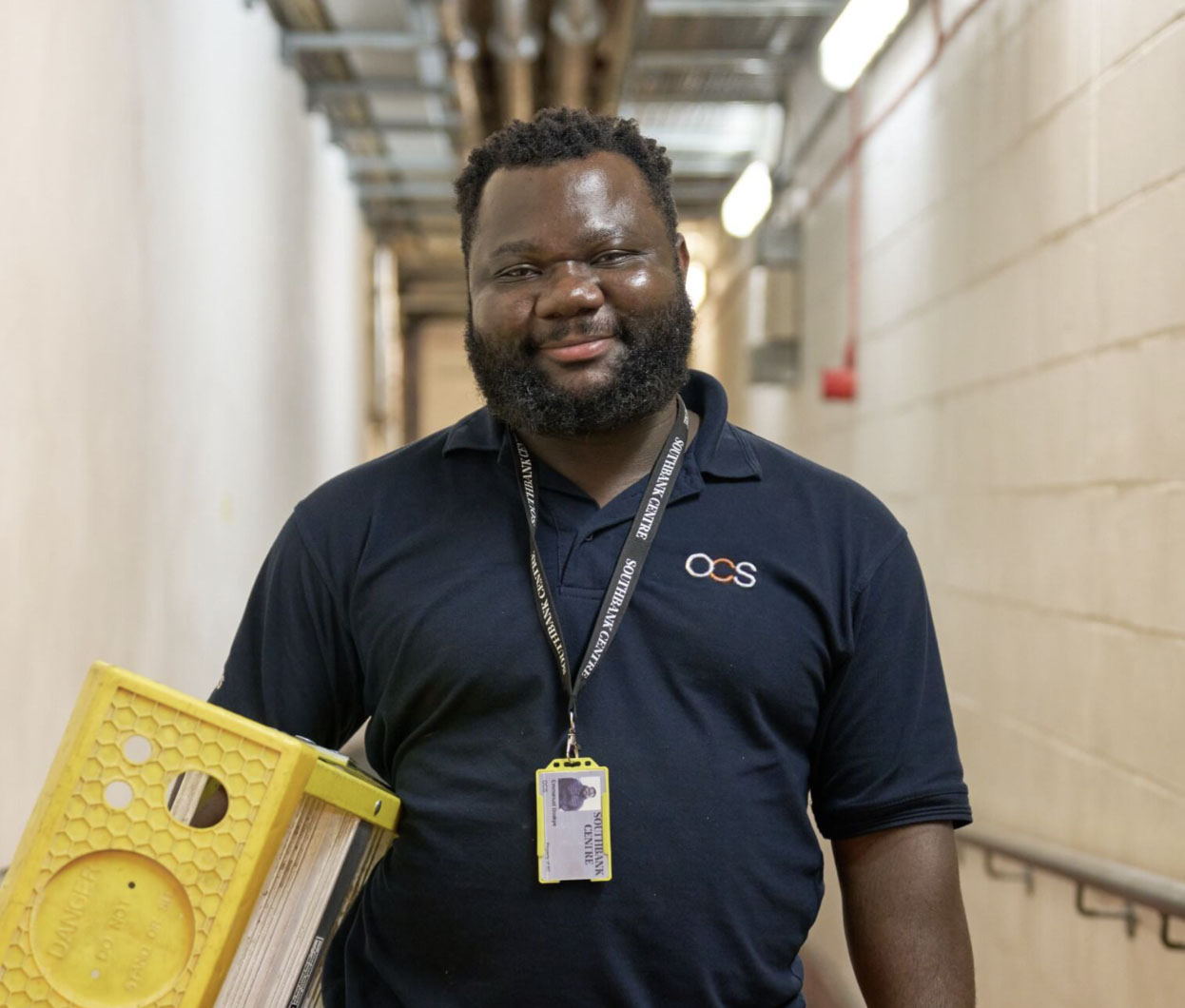 A smiling man with a beard, wearing a navy blue OCS polo shirt and lanyard, holds a yellow cleaning caddy in a corridor with cream brick walls and exposed pipes overhead.