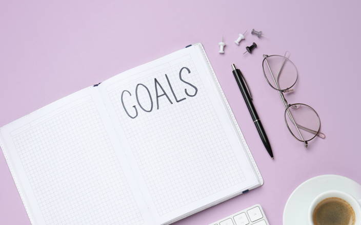 A notebook that says "goals" next to a cup of coffee, thumbtacks, a pen, and a pair of glasses for Confetti's Virtual Goal Setting Activities