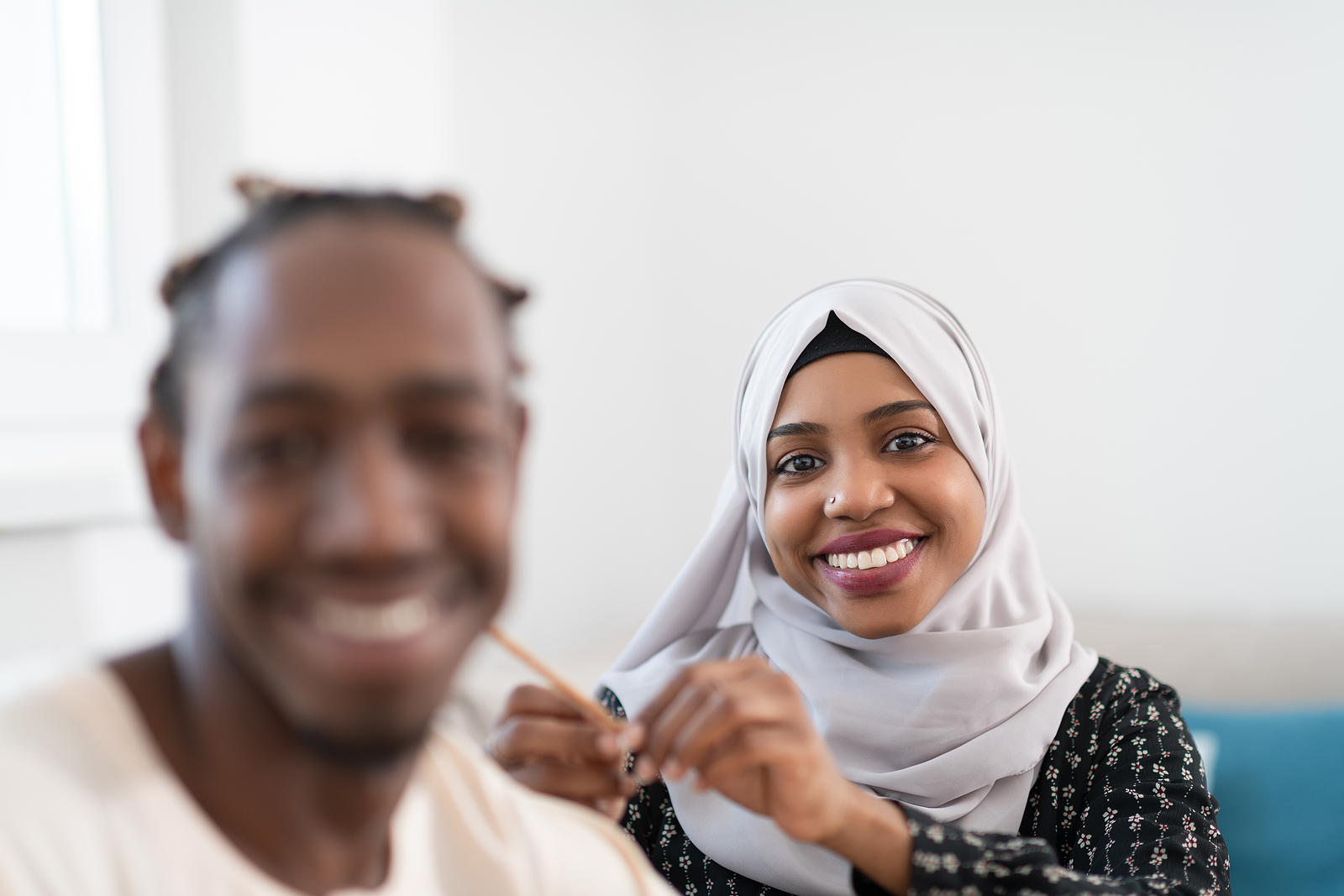 A woman in hijab, smiling and braiding a man's hair while he is looking into the camera as well and smiling.