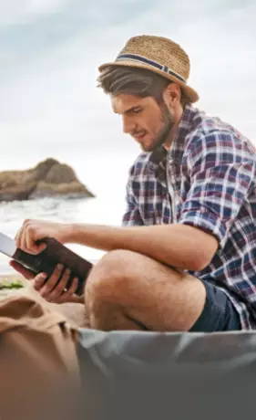 man in shorts, a plaid shirt and a straw hat, reading a book on the beach