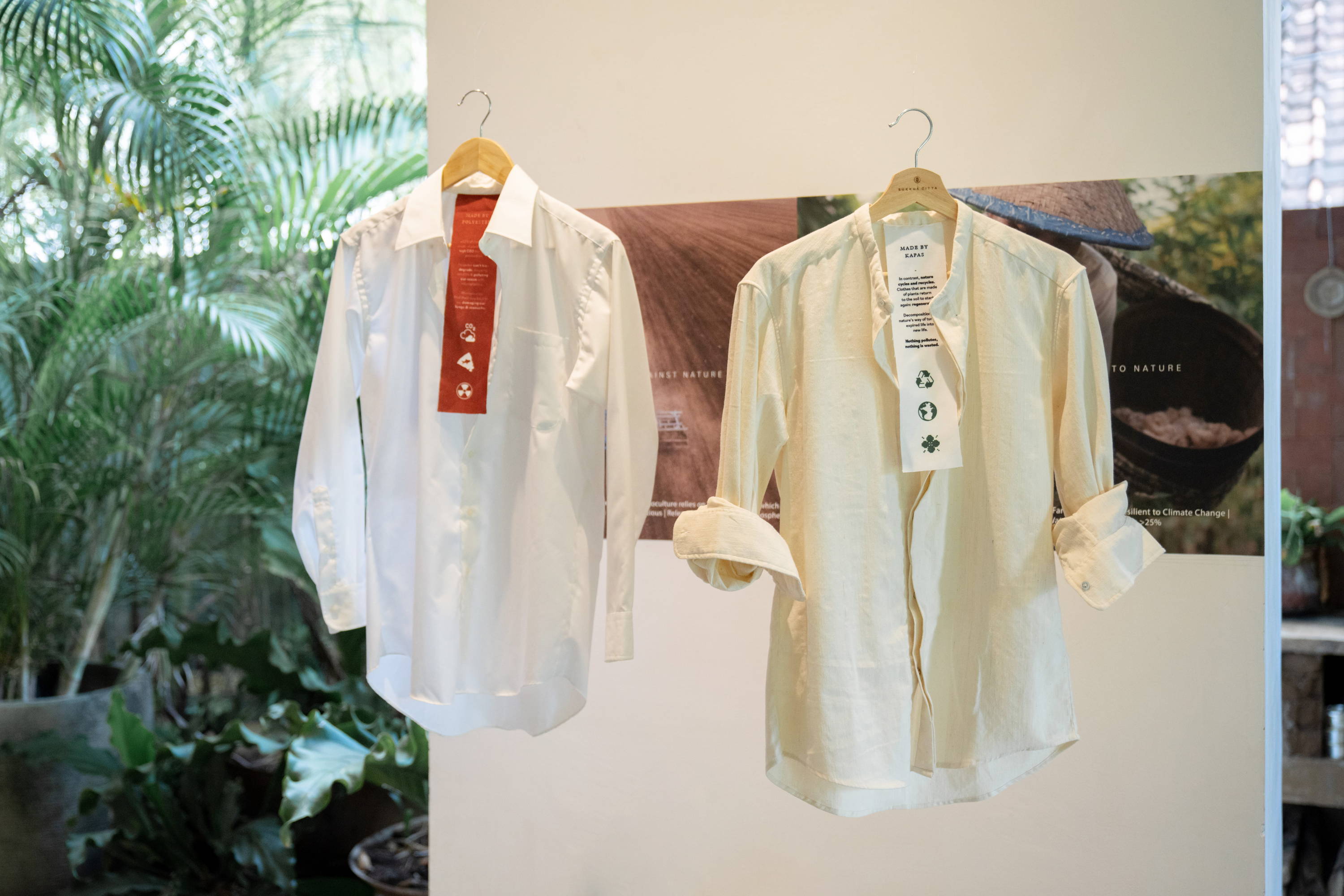 KAPAS Installation by SukkhaCitta in John Hardy Boutique & Gallery at Seminyak. A white polyester dress shirt with a red tag and a white regenerative cotton shirt with a white tag exhibited side by side. Text reads: More than 52% of clothes are made of plastic. Emits high carbon dioxide during its production. Polyester can't biodegrade, clogging landfills and polluting our ocean with microplastics. Microplastics find their way back to us, damaging our lungs and stomachs. In contrast, nature cycles and recycles. Clothes that are made of plants return to the soil to start again: regenerates. Decomposition is nature's way of turning expired life into new life. Nothing pollutes, nothing is wasted.