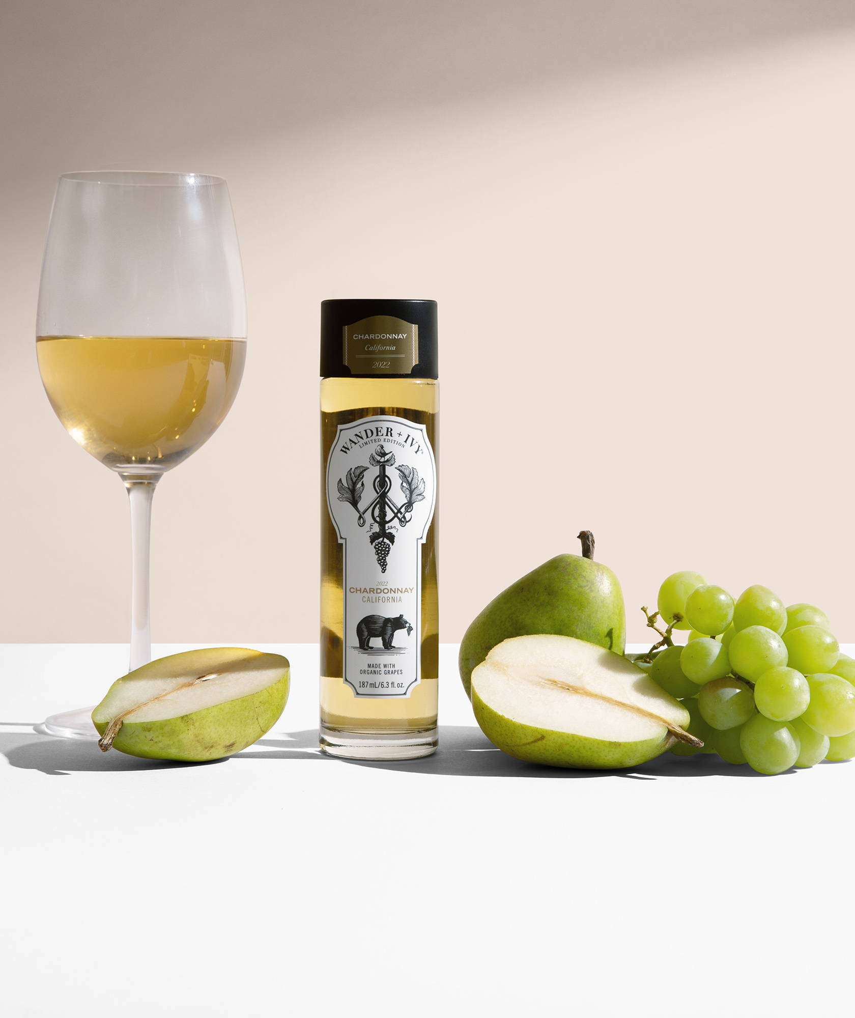 A single-serve bottle of Chardonnay next to a halved pear, bunch of white grapes, and a filled wine glass