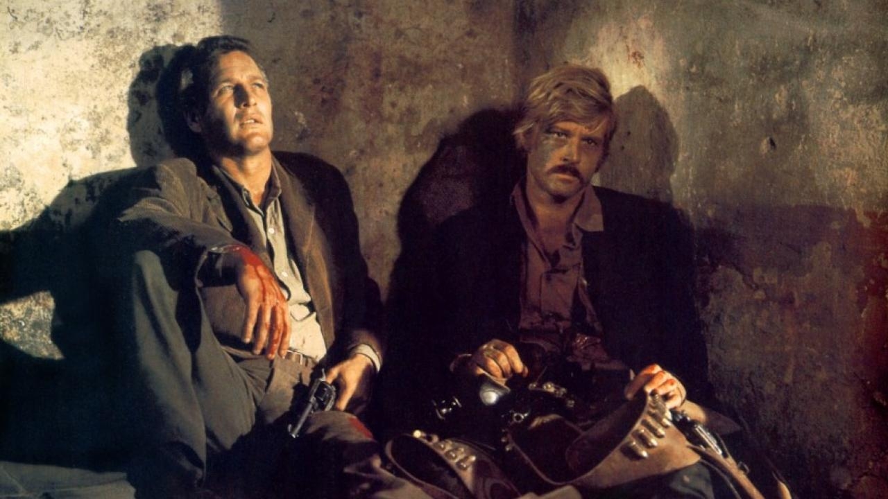 Butch cassidy and the sundance kid leaning on the wall on the ground, bloodied and holding weapons.
