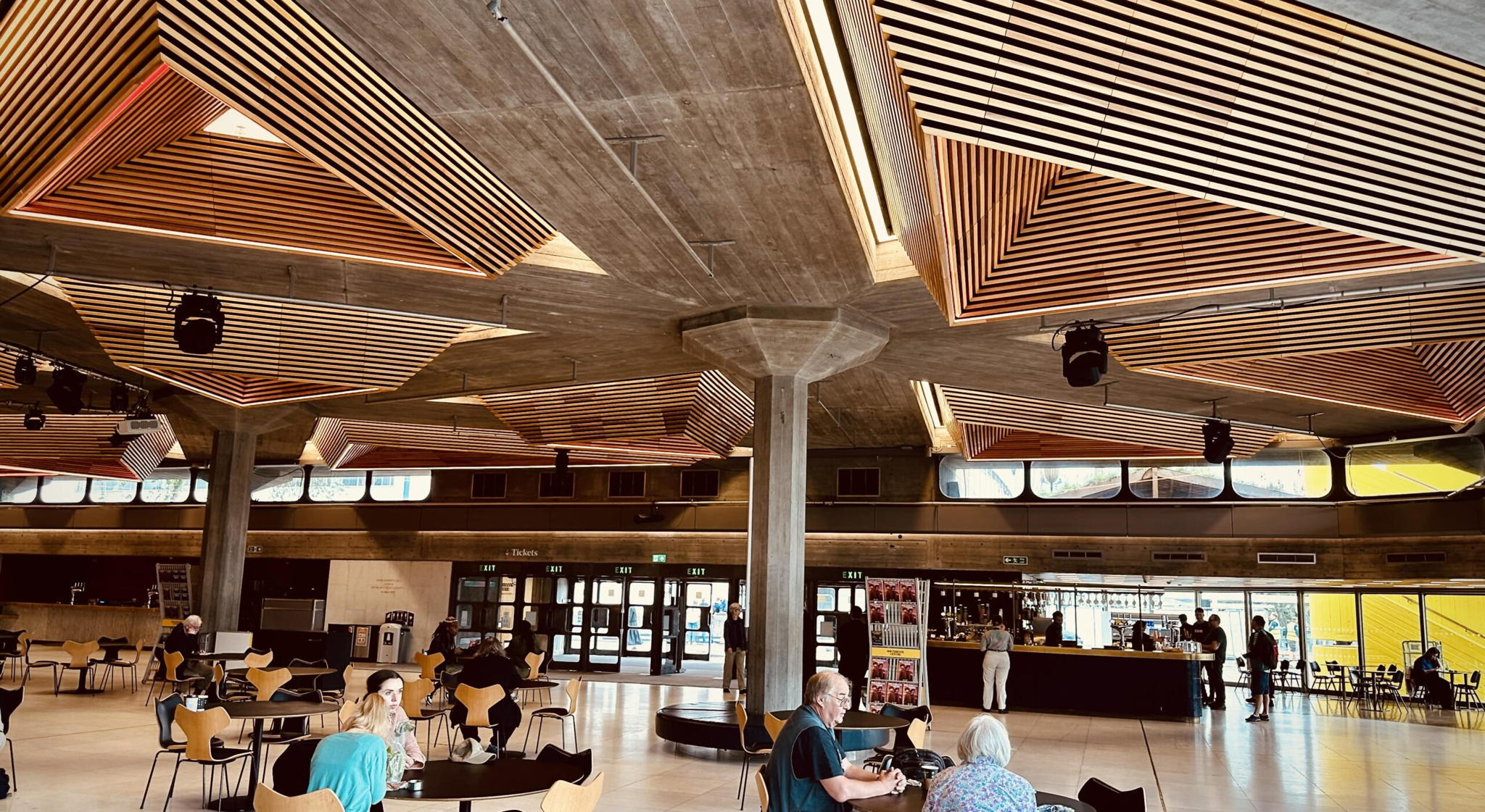 A spacious modern interior with geometric wooden ceiling panels, scattered tables and chairs, people sitting and chatting, and a bar area in the back. Large windows let in natural light.