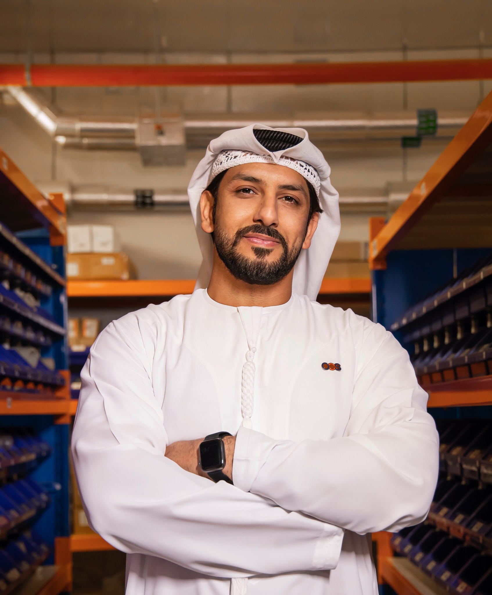 A man wearing a traditional white kandura and headscarf stands with arms crossed in a warehouse aisle, surrounded by shelves of organized boxes and packages.