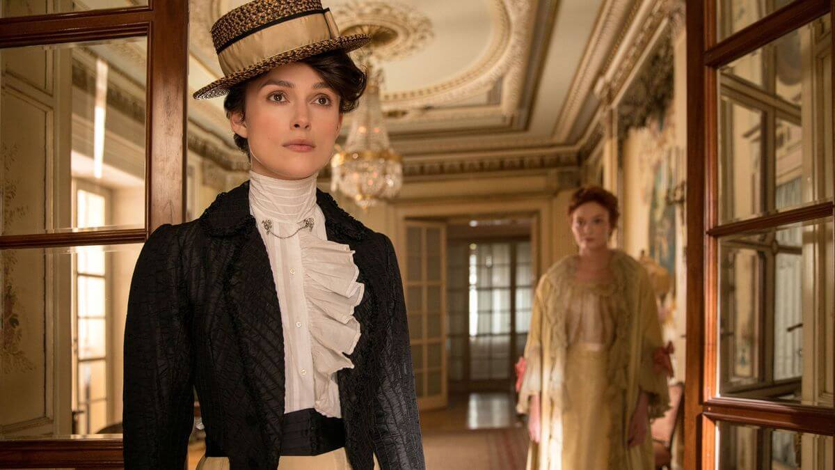 Colette dressed in the clothing from the time, walking and looking straight ahead inside a beautiful house.