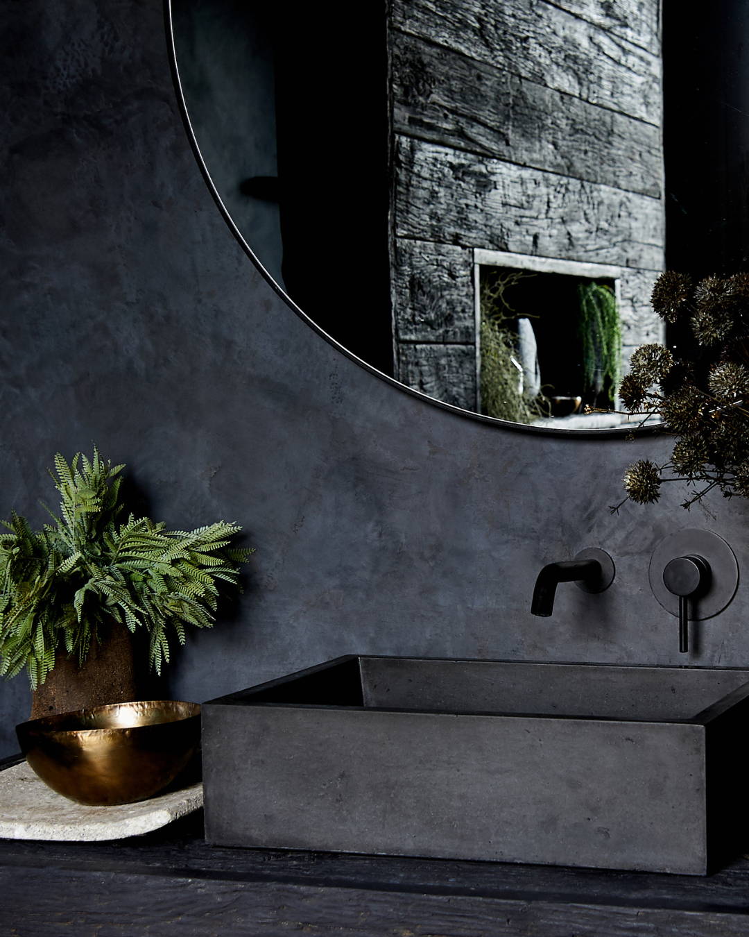A dark bathroom with black tadelakt walls, a large round mirror sits above a black concrete rectangular sink .which is next to an artificial plants and stems of mimosa.